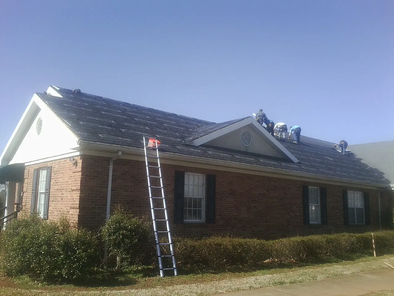 Crew tearing shingles off a brick ranch roof with a ladder set — Metal Roof Repair work by our crew in Wilber