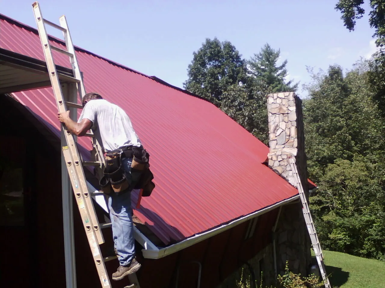Roofer on a ladder beside a red metal roof and stone chimney — Soffit & Fascia Repair by Roofing Brothers in Wilber